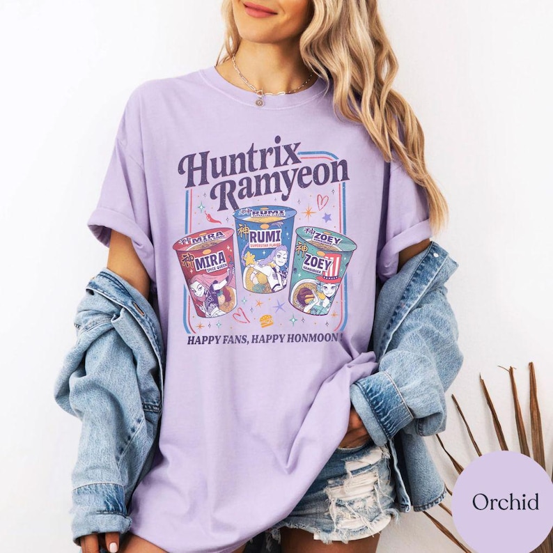 Puede incluir: Una camiseta de color lila claro con la inscripci&oacute;n "Huntrix Ramyeon" y dibujos de tres tazas de ramen. Cada taza lleva un nombre diferente: Mira, Rumi y Zoey. La frase "Happy Fans, Happy Honmoon!" est&aacute; impresa debajo.