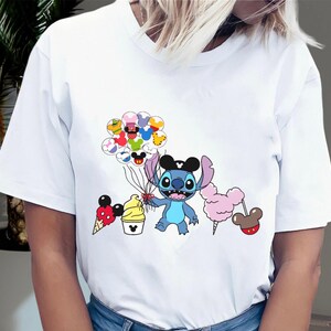 May include: White t-shirt with a blue Stitch character holding balloons with Disney characters on them. The balloons are red, yellow, green, blue, and purple. Stitch is wearing a black Mickey Mouse hat. There is a Mickey Mouse ice cream cone, a Mickey Mouse ice cream cup, and a pink cotton candy on sticks.