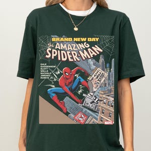 May include: Dark green t-shirt featuring a comic book cover design of Spider-Man. The graphic includes the text "BRAND NEW DAY" and "the AMAZING SPIDER-MAN". The superhero is depicted in a dynamic pose amidst a cityscape.