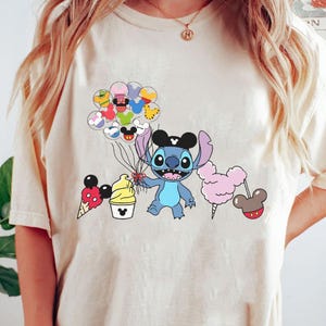 May include: A light beige t-shirt with a blue cartoon character holding colorful balloons. The character is wearing a black hat with a white star. The balloons are shaped like Mickey Mouse heads and have different colors and patterns.