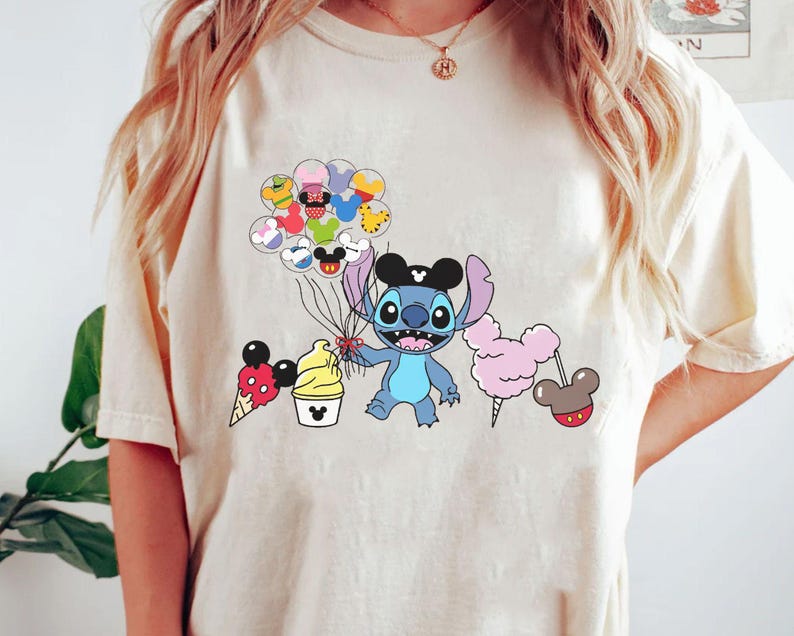 May include: A light beige t-shirt with a blue cartoon character holding colorful balloons. The character is wearing a black hat with a white star. The balloons are shaped like Mickey Mouse heads and have different colors and patterns.