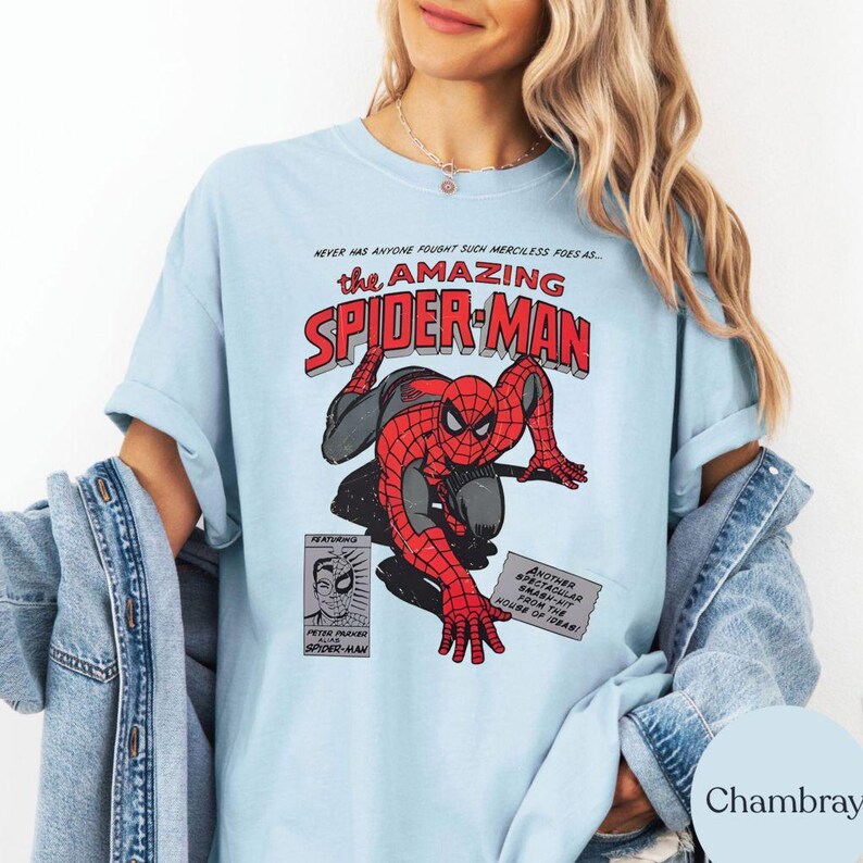 May include: Light blue t-shirt featuring a comic book graphic of Spider-Man in red and black. The text "the AMAZING SPIDER-MAN" is displayed above the image. Additional text includes "NEVER HAS ANYONE FOUGHT SUCH MERCILESS FOES AS..." and "FEATURING PETER PARKER SPIDER-MAN".