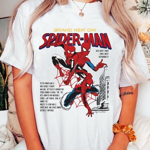 May include: White t-shirt featuring a comic book style graphic of Spider-Man. The design includes the words "BRAND NEW DAY" and "SPIDER-MAN" in red and blue, with text describing Spider-Man's origin story. The graphic is a vibrant illustration.