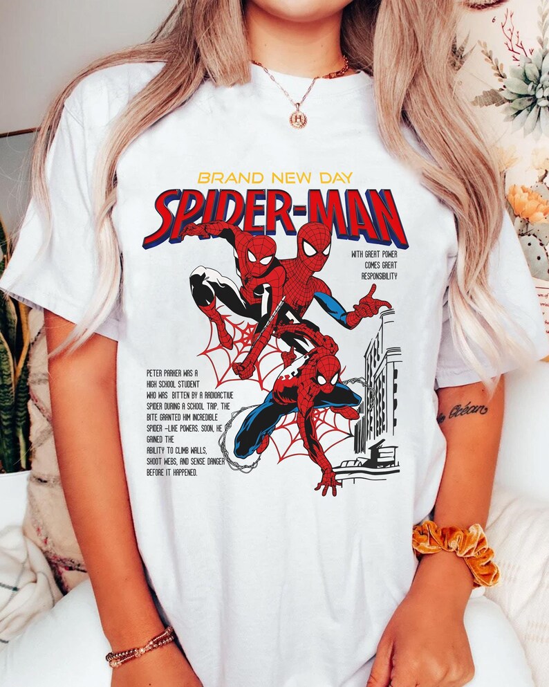 May include: White t-shirt featuring a comic book style graphic of Spider-Man. The design includes the words "BRAND NEW DAY" and "SPIDER-MAN" in red and blue, with text describing Spider-Man's origin story. The graphic is a vibrant illustration.
