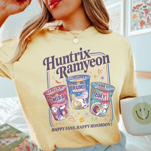 Puede incluir: Camiseta amarilla clara con el texto "Huntrix Ramyeon" en un dise&ntilde;o retro. El dise&ntilde;o incluye tres ilustraciones de tazas de ramen con los nombres "Mira", "Rumi" y "Zoey". La camiseta tambi&eacute;n tiene el texto "Happy Fans, Happy Honmoon!"