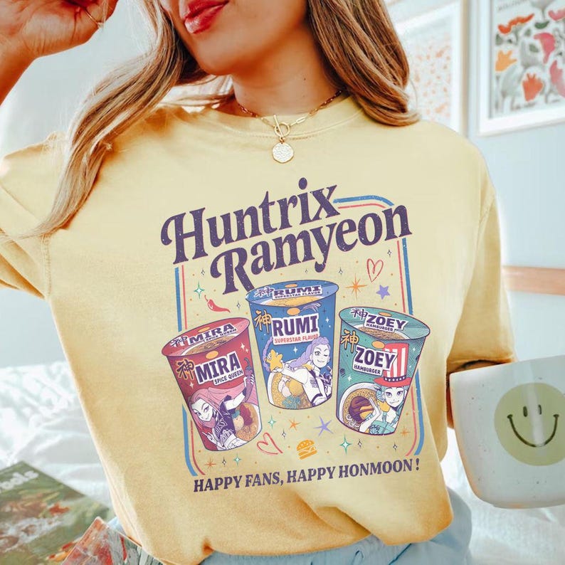 Puede incluir: Camiseta amarilla clara con el texto "Huntrix Ramyeon" en un dise&ntilde;o retro. El dise&ntilde;o incluye tres ilustraciones de tazas de ramen con los nombres "Mira", "Rumi" y "Zoey". La camiseta tambi&eacute;n tiene el texto "Happy Fans, Happy Honmoon!"
