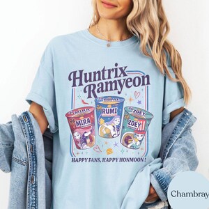 Puede incluir: Camiseta azul claro con el texto "Huntrix Ramyeon" e ilustraciones de tres tazas de ramen. Cada taza presenta un personaje y un sabor diferentes. La camiseta tambi&eacute;n tiene el texto "Happy Fans, Happy Honmoon!"