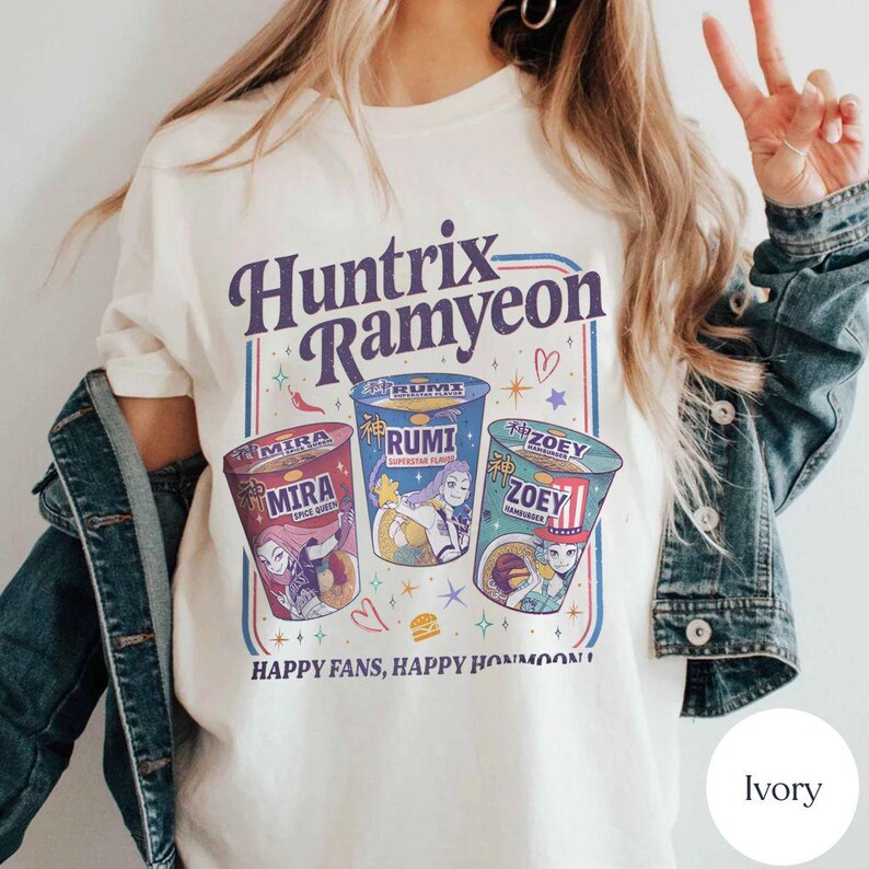 Puede incluir: Camiseta de color marfil con el texto "Huntrix Ramyeon" y ilustraciones de tazas de ramen. Las tazas est&aacute;n etiquetadas como "Mira", "Rumi" y "Zoey". El dise&ntilde;o incluye estrellas, corazones y la frase "Happy Fans, Happy Honmoon!"