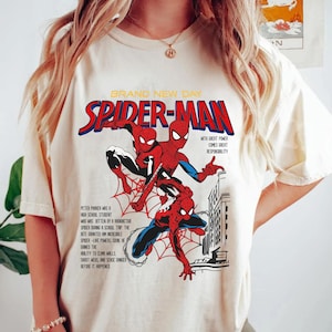 May include: Cream-colored t-shirt featuring a comic book-style graphic of Spider-Man. The design includes the words "BRAND NEW DAY" and "SPIDER-MAN" in red and blue, along with text describing Spider-Man's origin and powers.