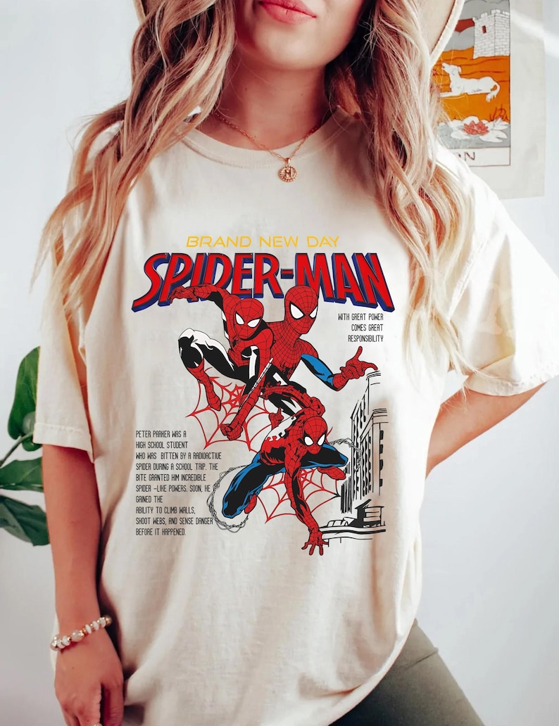 May include: Cream-colored t-shirt featuring a comic book-style graphic of Spider-Man. The design includes the words "BRAND NEW DAY" and "SPIDER-MAN" in red and blue, along with text describing Spider-Man's origin and powers.