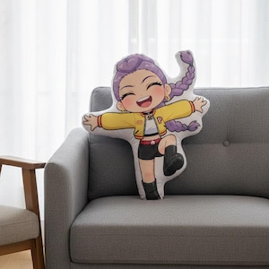 May include: A cushion shaped like a cartoon character with purple braided hair, a yellow jacket, black shorts, and black boots. The cushion is placed on a grey sofa.