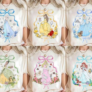 Può includere: Sei magliette color crema con diversi disegni di principesse. Ogni maglietta presenta un'illustrazione colorata di una principessa all'interno di una cornice decorativa. Le principesse includono Elsa, Belle, Cenerentola, Tiana, Aurora e Ariel.
