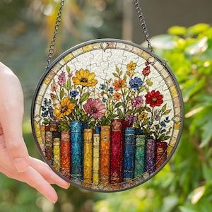 Peut inclure: Vitrail rond représentant un arrangement floral coloré au-dessus d'une rangée de livres. Les fleurs sont jaunes, roses, rouges et bleues. Les livres sont de couleurs variées. Une chaîne noire est attachée pour l'accrocher.