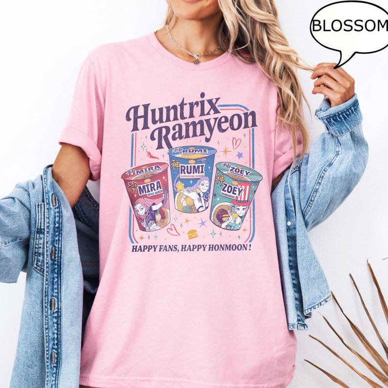 Puede incluir: Camiseta rosa claro con la inscripci&oacute;n "Huntrix Ramyeon" y tres ilustraciones de vasos de ramen. Cada vaso lleva un nombre diferente: Mira, Rumi y Zoey. La camiseta tambi&eacute;n incluye el texto "Happy Fans, Happy Honmoon!"
