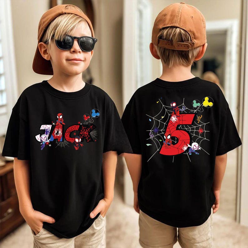 Pu&ograve; includere: T-shirt nera a tema Spider-Man. La parte anteriore presenta il nome "JAC" con grafica di Spider-Man. Il retro mostra un grande numero "5" rosso circondato da una ragnatela e personaggi di Spider-Man.