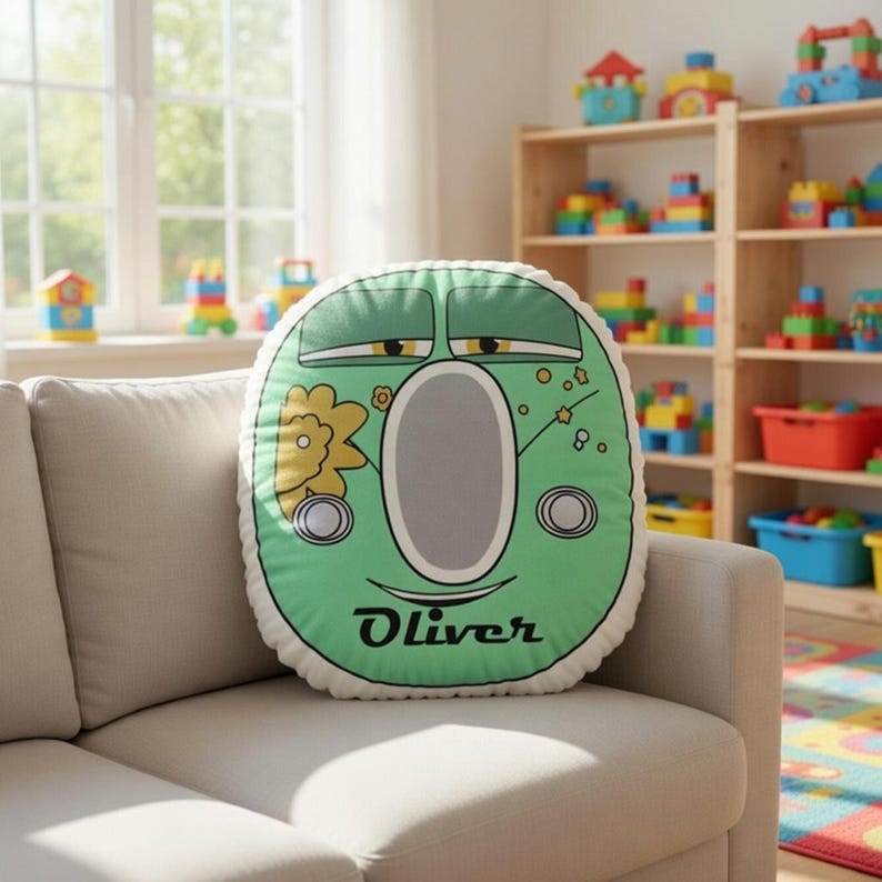 May include: A round, green decorative pillow with a cartoon face design, featuring a large oval mouth and two circular eyes. The pillow has the name "Oliver" printed in a script font. The pillow is resting on a beige sofa in a playroom setting.