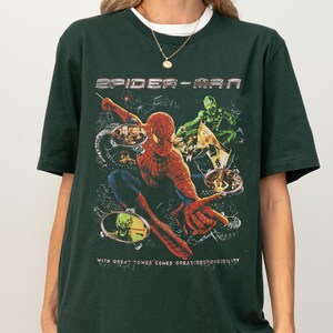 Puede incluir: Camiseta verde oscuro con un dise&ntilde;o de Spider-Man. El dise&ntilde;o muestra a Spider-Man en acci&oacute;n, con las palabras "SPIDER-MAN" arriba y la frase "WITH GREAT POWER COMES GREAT RESPONSIBILITY" abajo. El dise&ntilde;o tambi&eacute;n presenta al Duende Verde.
