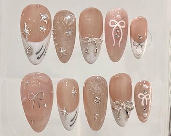 May include: Ten nude-colored fake nails with various white and silver designs. The designs include bows, stars, and swirls.