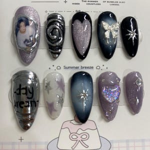May include: A collection of ten artificial nails with diverse designs. The nails showcase silver, black, and purple hues, adorned with glitter, stars, and a bow. Some nails have text or images. The text "Summer's Whimsy" is at the top of the image.