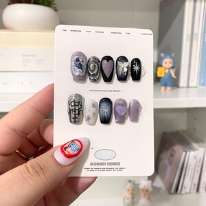 May include: A collection of false nails with diverse designs, such as a swirling silver pattern, a purple heart, and a black nail with a white bow. The nails are presented on a white card with the text "SUMMER DESIRE".