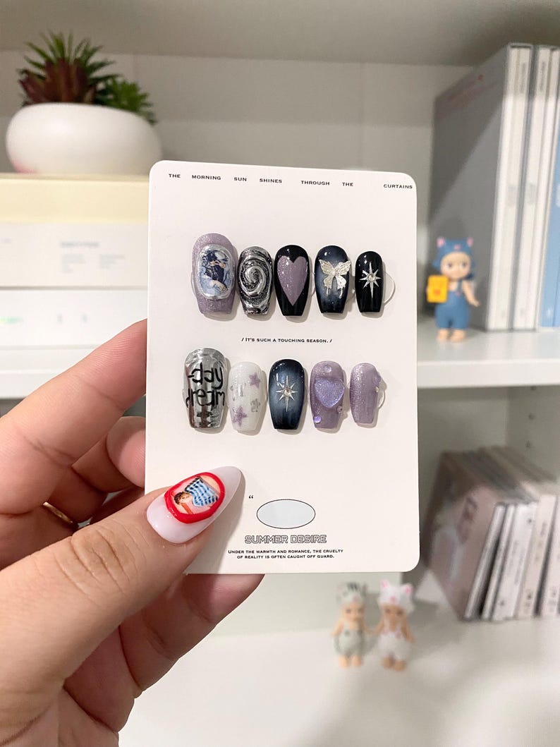 May include: A collection of false nails with diverse designs, such as a swirling silver pattern, a purple heart, and a black nail with a white bow. The nails are presented on a white card with the text "SUMMER DESIRE".