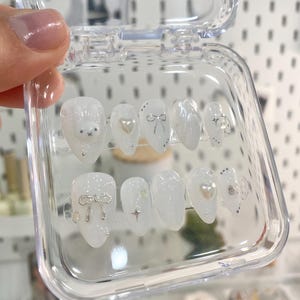 May include: A clear plastic box containing ten white press-on nails with various silver and pearl embellishments, including bows, stars, and hearts.