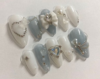 May include: Set of ten press-on nails with a white and light blue design. The nails feature various embellishments, including rhinestones, pearls, chains, and bows.