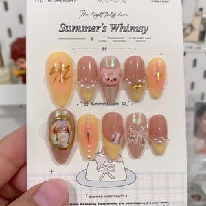May include: A collection of false nails with diverse designs. Some nails have a yellow and orange gradient with gold details. Others feature a cat face, pearls, or a photo. The card reads "Summer's Whimsy."