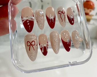 May include: A set of ten false nails with a red base and a white glitter tip. Each nail has a silver bow design.
