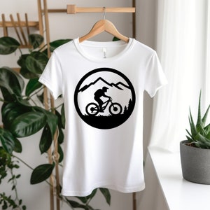 Puede incluir: Camiseta blanca con una silueta negra de un ciclista de monta&ntilde;a que recorre un paisaje monta&ntilde;oso en un c&iacute;rculo.