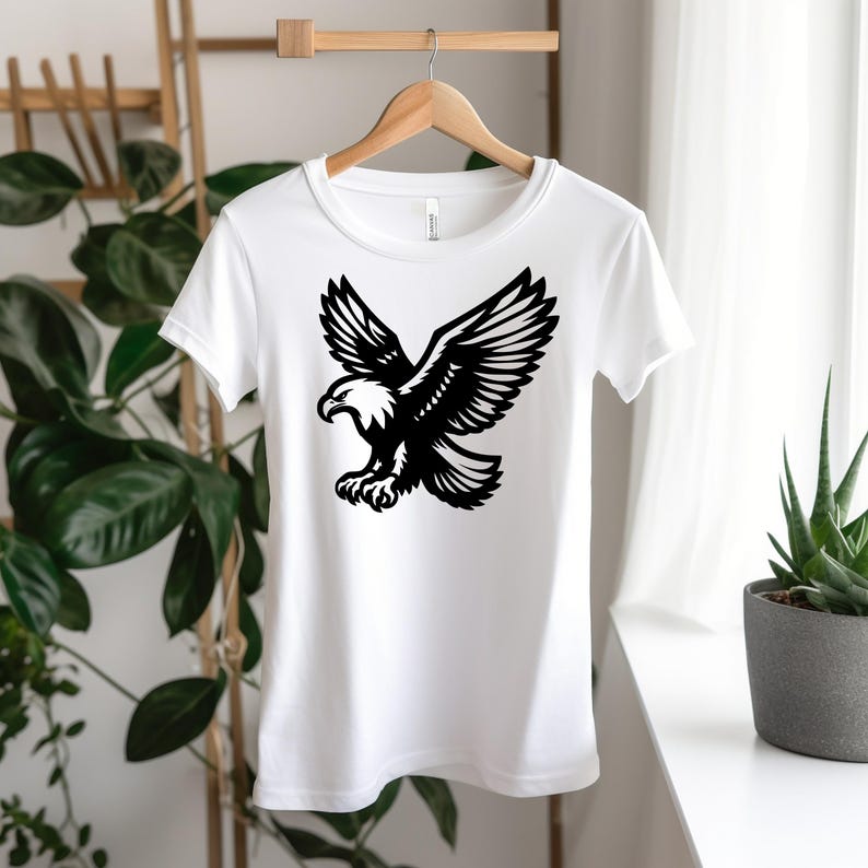 Puede incluir: Camiseta blanca con una silueta negra de un &aacute;guila calva con las alas extendidas.
