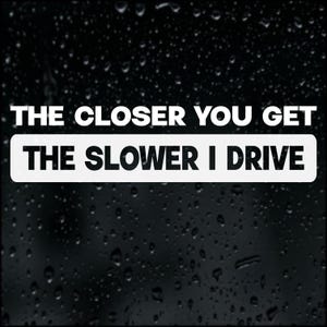 May include: A white rectangular sticker with the text "THE CLOSER YOU GET THE SLOWER I DRIVE" in a bold, sans-serif font. The background is black, possibly a car window, with water droplets.