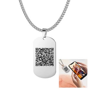 Peut inclure: Pendentif en forme de plaque d'identité argenté avec un code QR, suspendu à une chaîne assortie. La plaque est rectangulaire avec des coins arrondis. Un smartphone affiche une photo, et un petit pendentif est également visible.