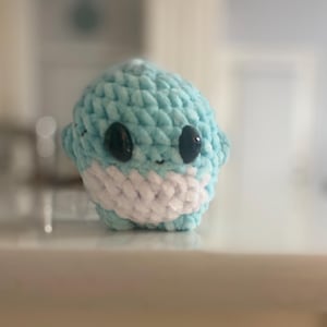 May include: A small, handmade crochet shark in shades of light blue and white. The shark has two black button eyes and small fins. The plush toy is soft and cuddly, perfect for children or as a decorative item.