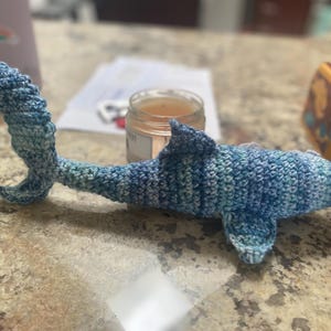 May include: A handmade crochet shark in shades of blue and white. The shark has a dorsal fin and a curved tail. The toy is lying on a speckled countertop.