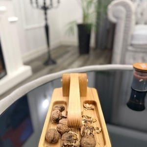 Può includere: Uno schiaccianoci in legno con un vassoio integrato, contenente noci e gusci. Lo schiaccianoci ha una leva e una base rettangolare con due scomparti. Il legno è di colore chiaro e il vassoio è pieno di noci intere e rotte.