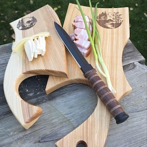 Puede incluir: Tres tablas de cortar de madera con un cuchillo y alimentos. Las tablas tienen una forma única con un asa y una escena de bosque decorativa. Queso en rodajas, tocino y cebolletas están dispuestos en las tablas. El cuchillo tiene una hoja negra y un mango de cuero marrón.
