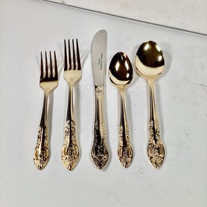 Puede incluir: Conjunto de cinco piezas de cubiertos chapados en oro, que incluyen dos tenedores, un cuchillo y dos cucharas. Los mangos tienen un diseño adornado.