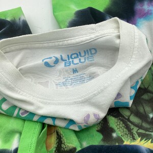 May include: White crew neck t-shirt with the "LIQUID BLUE" logo in blue. The shirt is a medium size and features a tie-dye design in green, blue, and purple. The shirt is made of cotton.