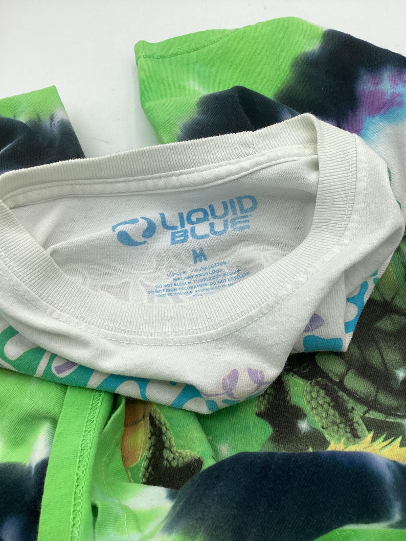 May include: White crew neck t-shirt with the "LIQUID BLUE" logo in blue. The shirt is a medium size and features a tie-dye design in green, blue, and purple. The shirt is made of cotton.