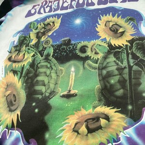 May include: A white t-shirt with a tie-dye design and the words "Grateful Dead" in a graphic. The graphic includes sunflowers, turtles, a candle, and a night sky scene. The shirt has a crew neck.