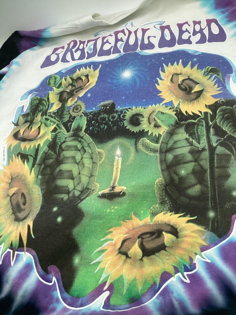 May include: A white t-shirt with a tie-dye design and the words "Grateful Dead" in a graphic. The graphic includes sunflowers, turtles, a candle, and a night sky scene. The shirt has a crew neck.