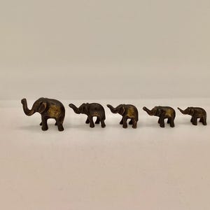 May include: Five small, bronze-coloured elephant figurines are displayed in a row. The elephants diminish in size from left to right. Each figurine has a trunk, ears, and legs. The elephants are on a plain, off-white surface.