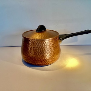 May include: A hammered copper saucepan with a black handle and lid. The saucepan has a rounded shape and a textured surface.