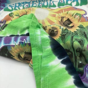 May include: A white tie-dye t-shirt with the words "Grateful Dead" in green. The shirt features a colourful graphic of sunflowers and a sun. The sleeves are folded, showing a green tie-dye pattern.