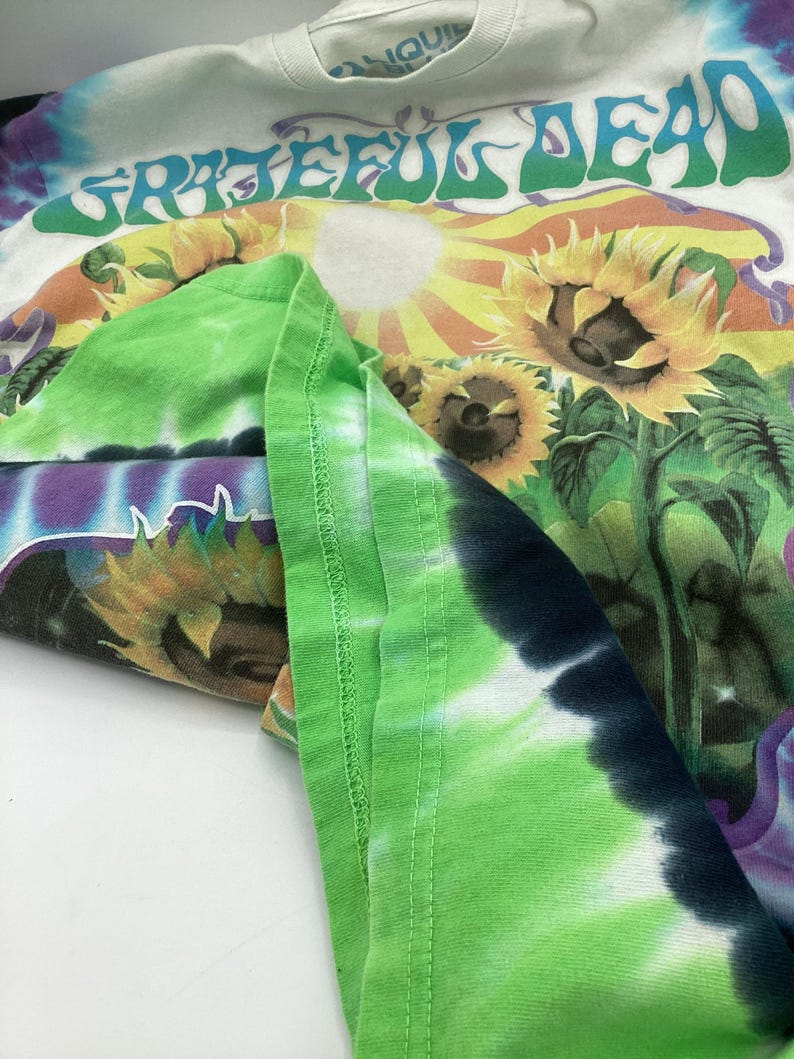 May include: A white tie-dye t-shirt with the words "Grateful Dead" in green. The shirt features a colourful graphic of sunflowers and a sun. The sleeves are folded, showing a green tie-dye pattern.