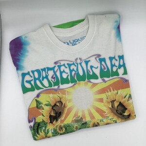 May include: White t-shirt with a tie-dye pattern on the shoulders and the text "Grateful Dead" in green and purple. The shirt features a graphic of sunflowers and a sun.