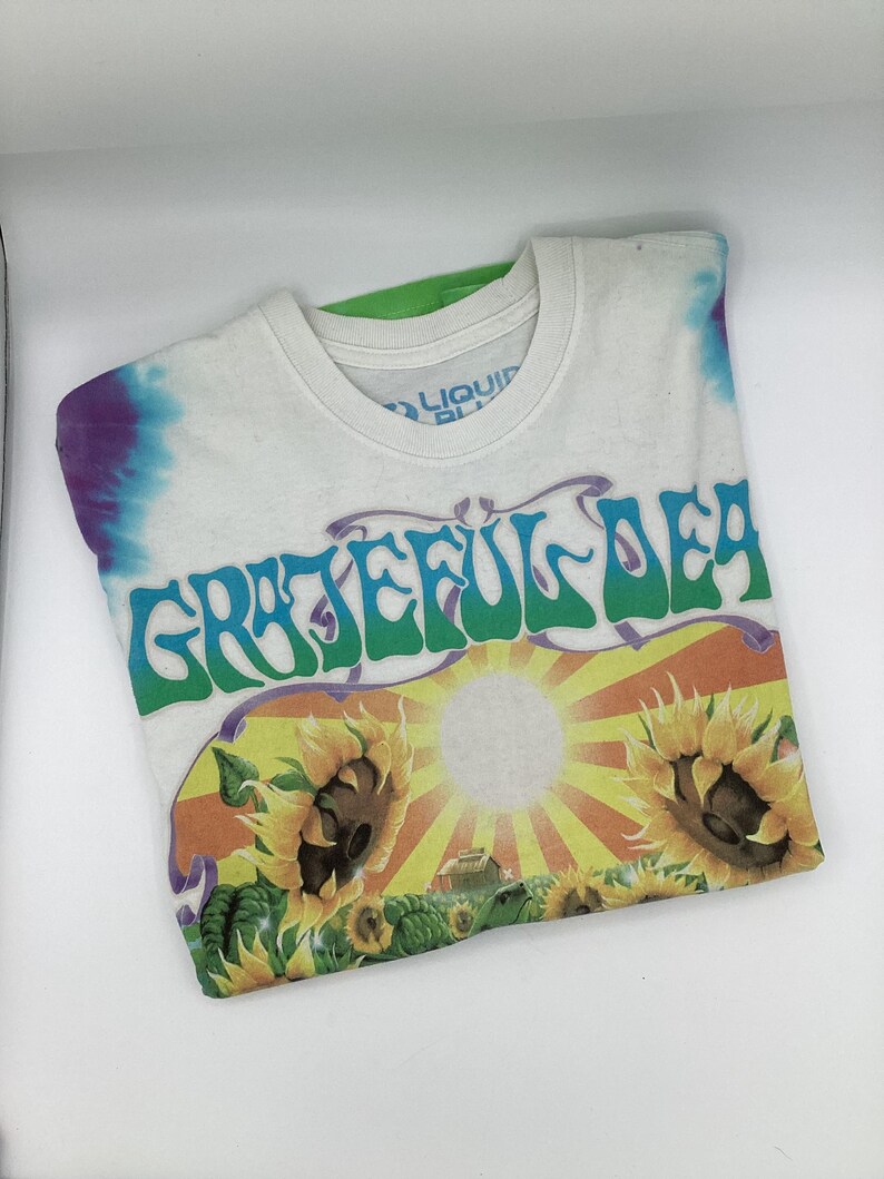 May include: White t-shirt with a tie-dye pattern on the shoulders and the text "Grateful Dead" in green and purple. The shirt features a graphic of sunflowers and a sun.