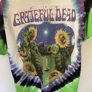 May include: White tie-dye t-shirt with the words "Grateful Dead" in purple. The graphic shows sunflowers, turtles, and a candle against a starry night. The shirt's edges are dyed in green, purple, and black, creating a vibrant look.