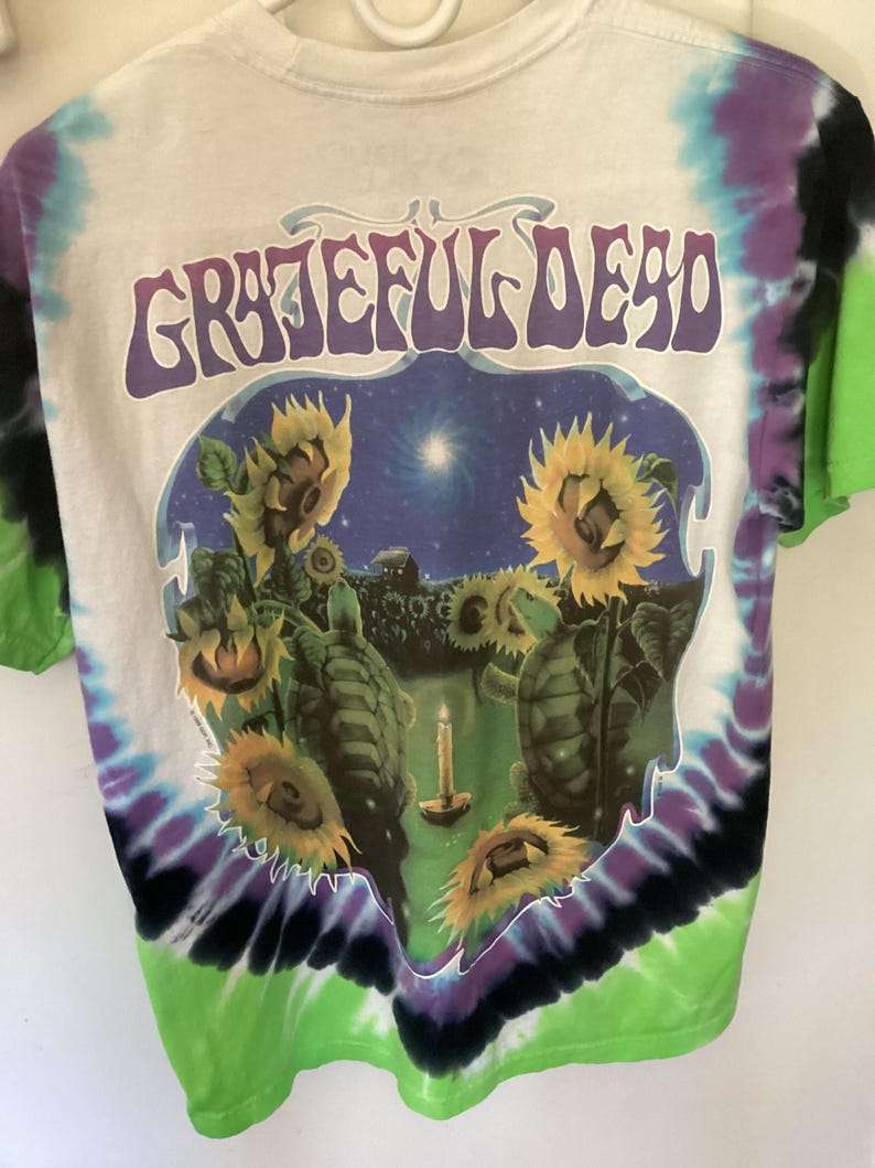 May include: White tie-dye t-shirt with the words "Grateful Dead" in purple. The graphic shows sunflowers, turtles, and a candle against a starry night. The shirt's edges are dyed in green, purple, and black, creating a vibrant look.